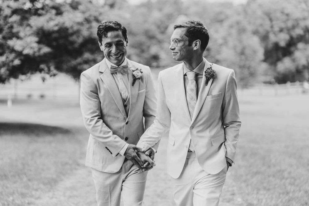 Two grooms in matching light gray suits smile and hold hands outdoors on their wedding day in a black and white photo.