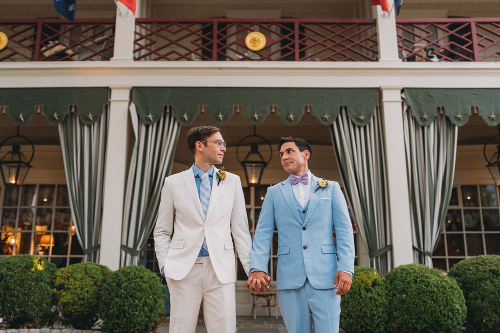 Two grooms in suits holding hands outside a historic venue with green awnings and flags.
