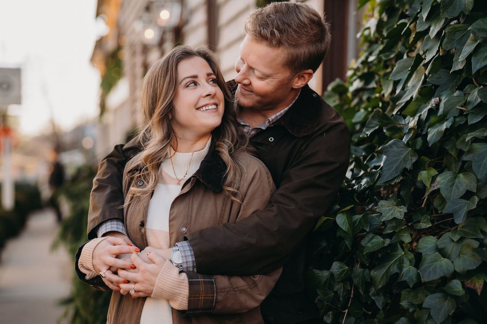 Couple embracing outdoors near ivy-covered wall, smiling warmly in autumn jackets on a charming city street.