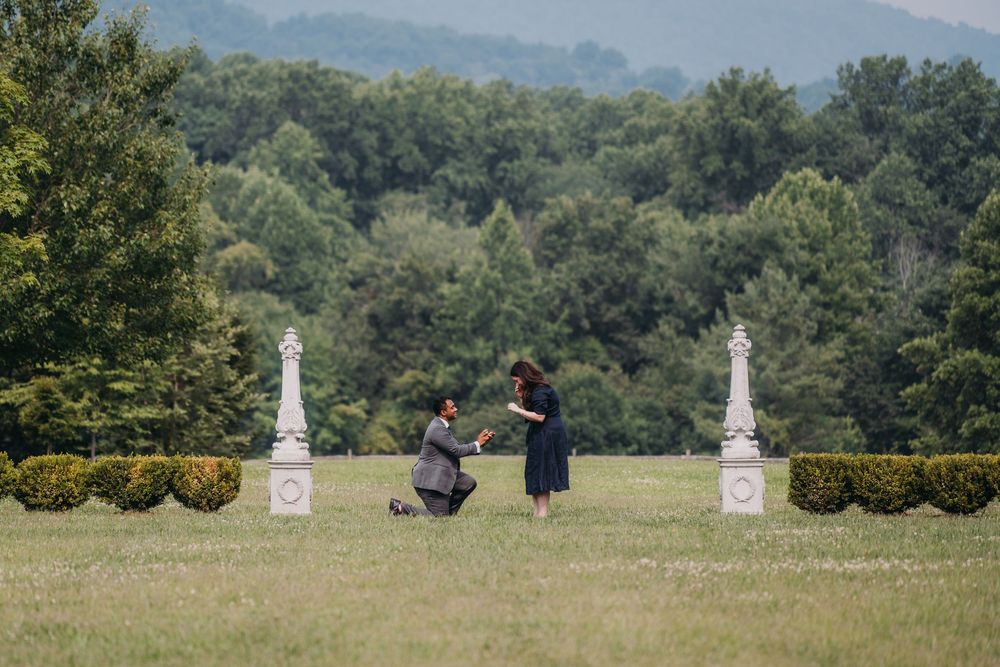 Man kneeling on grass proposing to woman outdoors, with white stone pillars and lush green trees in background.