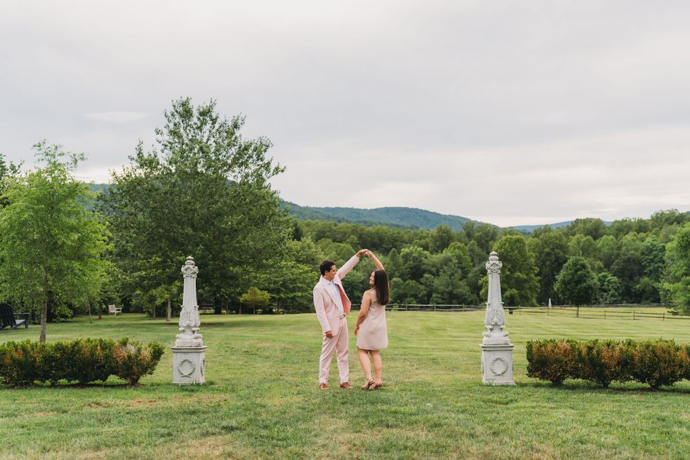 Couple dancing on lush green lawn with stone pillars and mountain backdrop on a cloudy day.