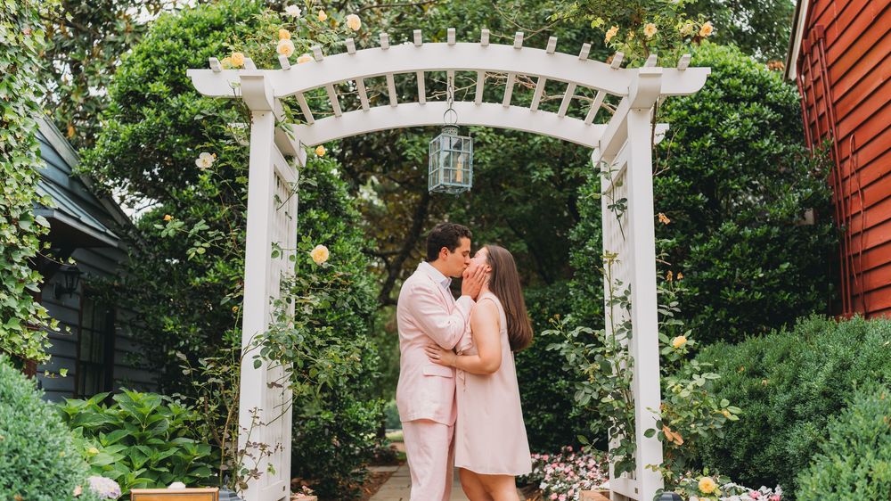 Couple sharing a romantic kiss under a white garden arbor covered in lush greenery and flowers.