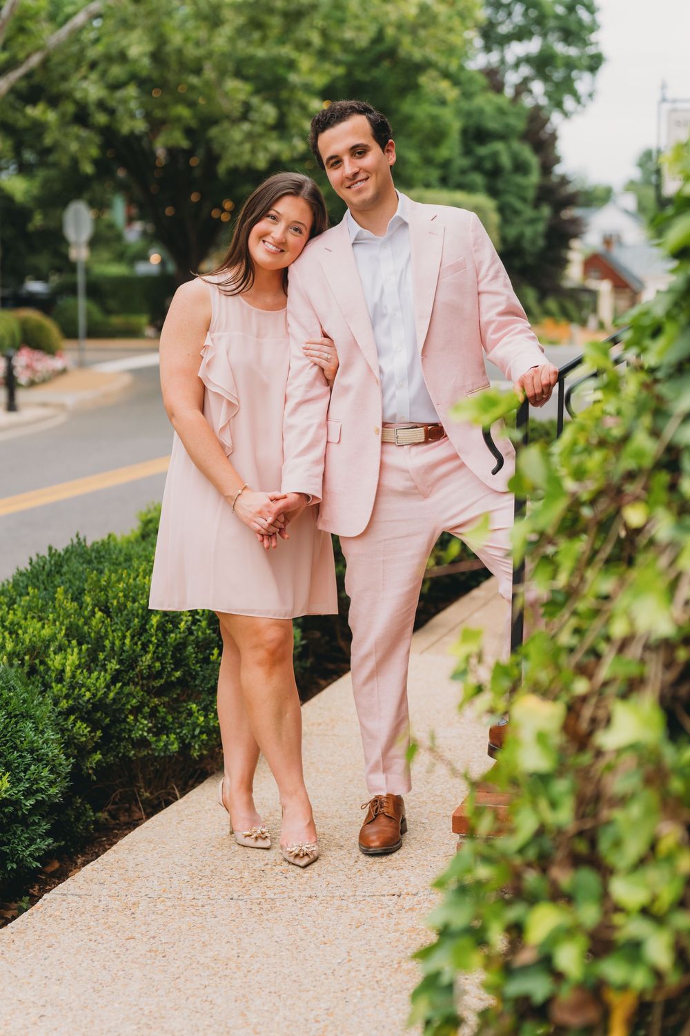 Couple in matching pink outfits posing on a sidewalk surrounded by lush greenery on a sunny day.