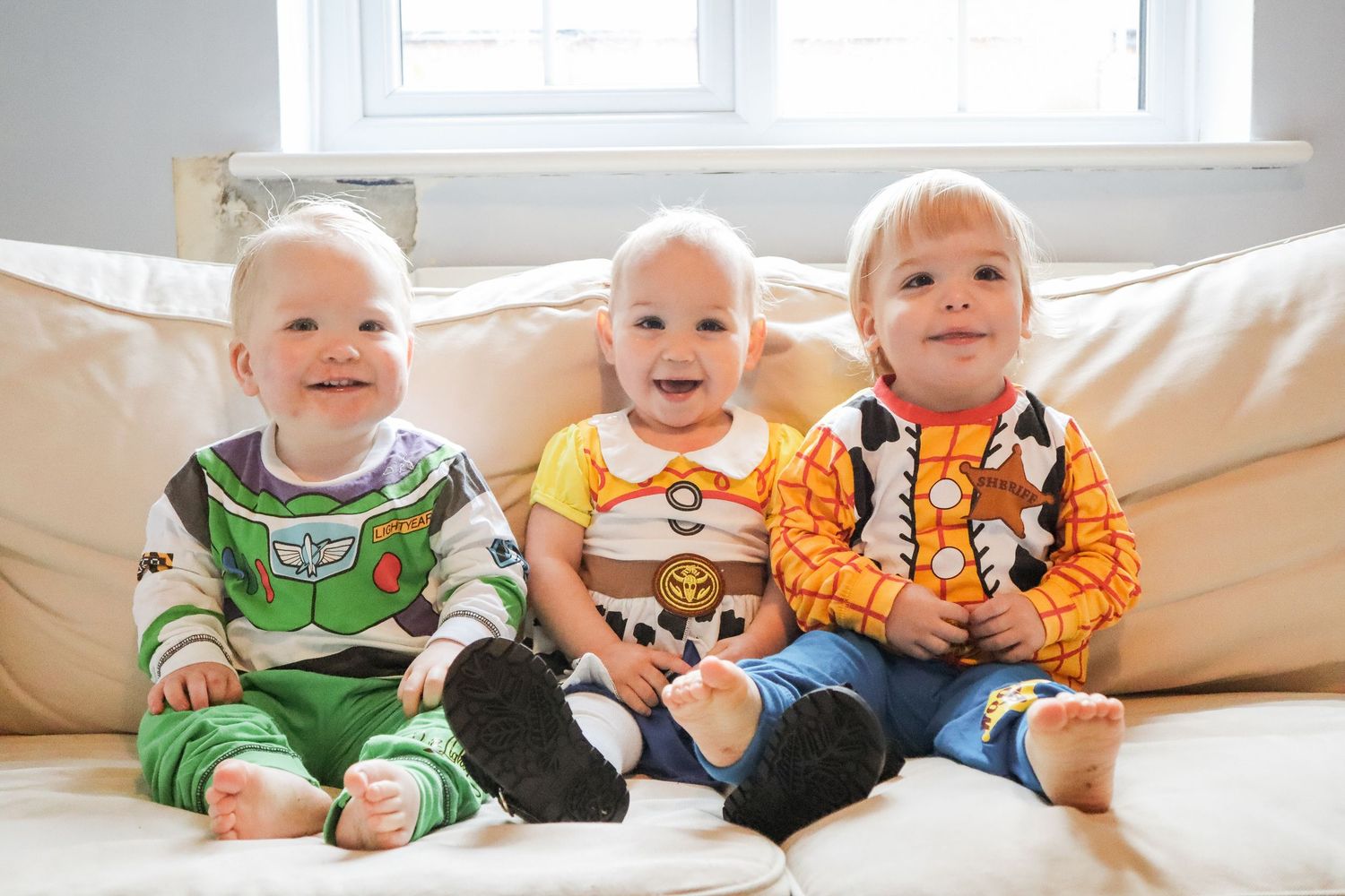 Three toddlers dressed in Toy Story costumes as Buzz Lightyear, Woody, and Jessie sitting on a beige sofa.