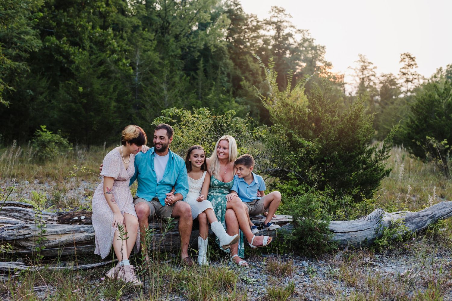 A happy family of five sits together on a fallen log in a lush green outdoor setting during golden hour.