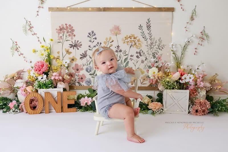 Baby's first birthday photo with floral backdrop, wooden ONE letters, and infant in blue romper seated among pink and yellow flowers.
