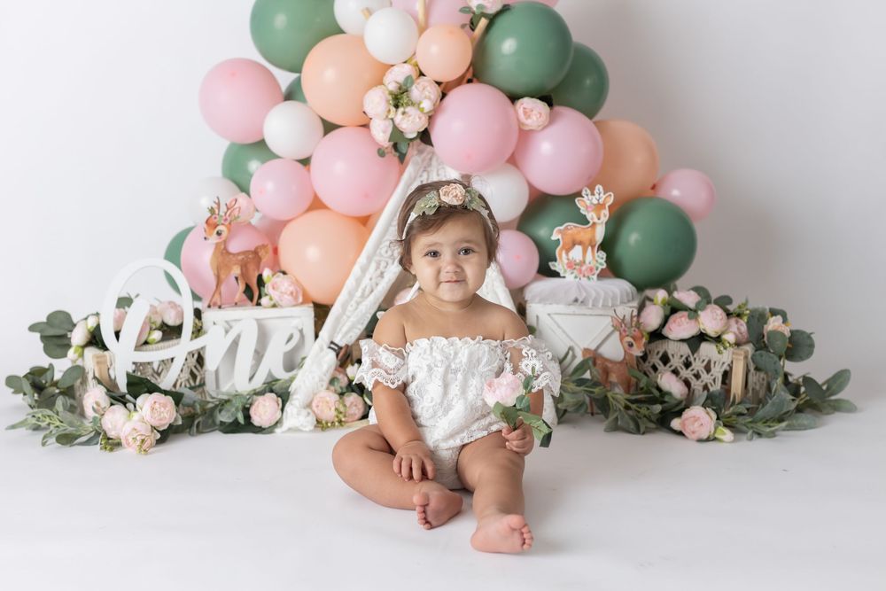 Baby girl in white romper sits before colorful balloon arch with florals and deer decor for first birthday cake smash.