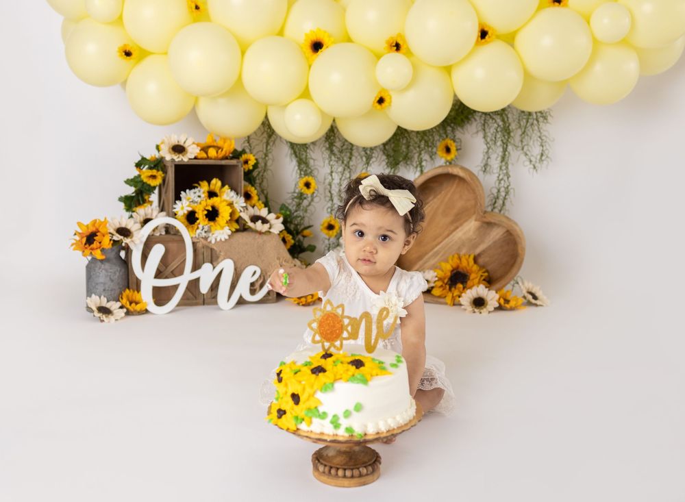 Baby girl's first birthday cake smash with sunflower theme, yellow balloons, and "One" sign backdrop.