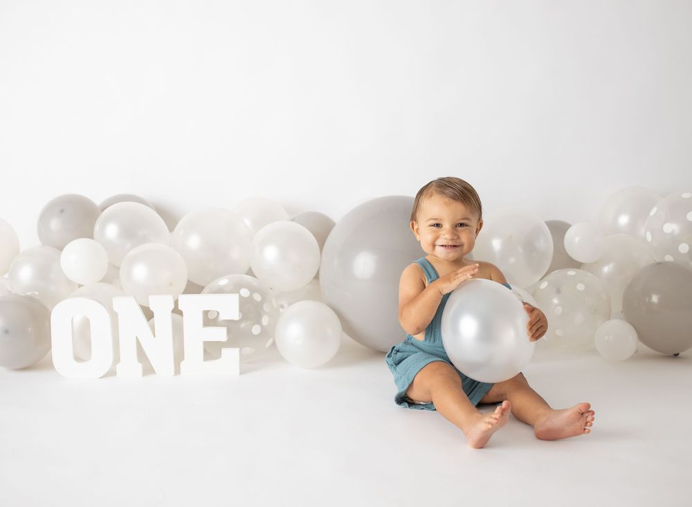 A smiling baby celebrates their first birthday sitting among white and silver balloons with large ONE letters on a white backdrop.