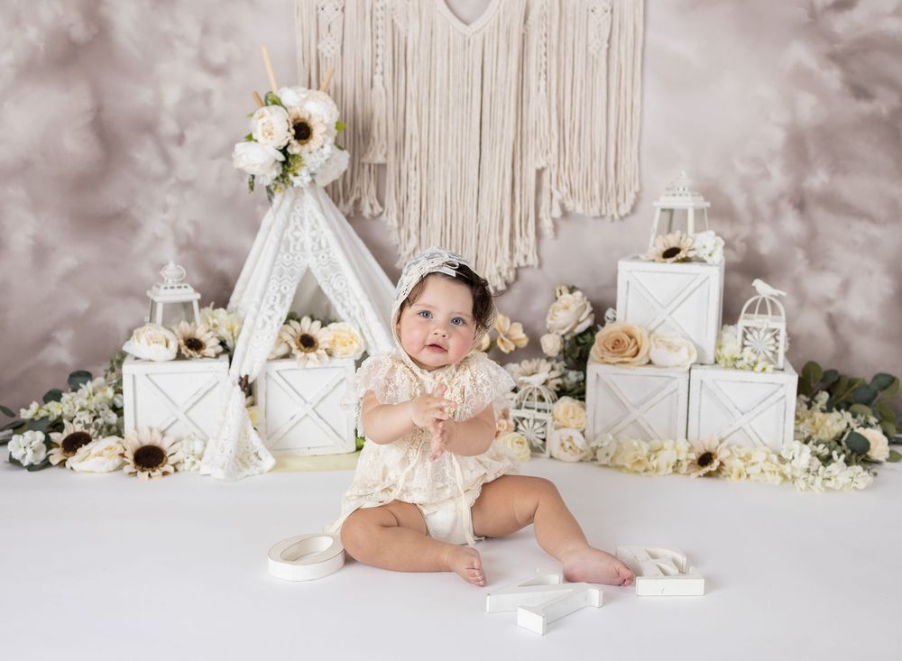 Baby girl in lace romper sits before boho teepee backdrop with white boxes, lanterns, and sunflower floral arrangements.
