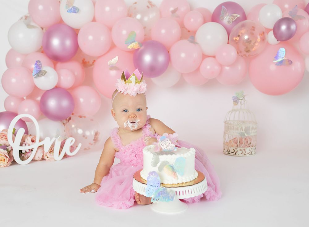 Baby girl in pink tutu celebrates first birthday with unicorn cake surrounded by pink balloon garland.