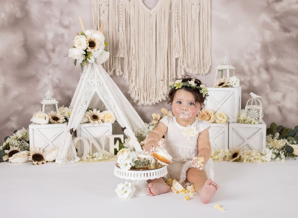 Baby girl in white romper enjoys first birthday cake smash with boho teepee backdrop and floral decorations.