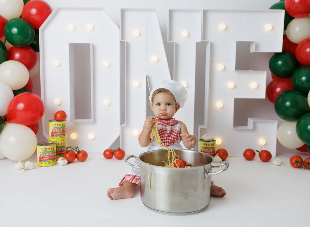 Baby in chef hat sits in large pot with spaghetti, surrounded by tomatoes and balloons for ONE birthday photo.