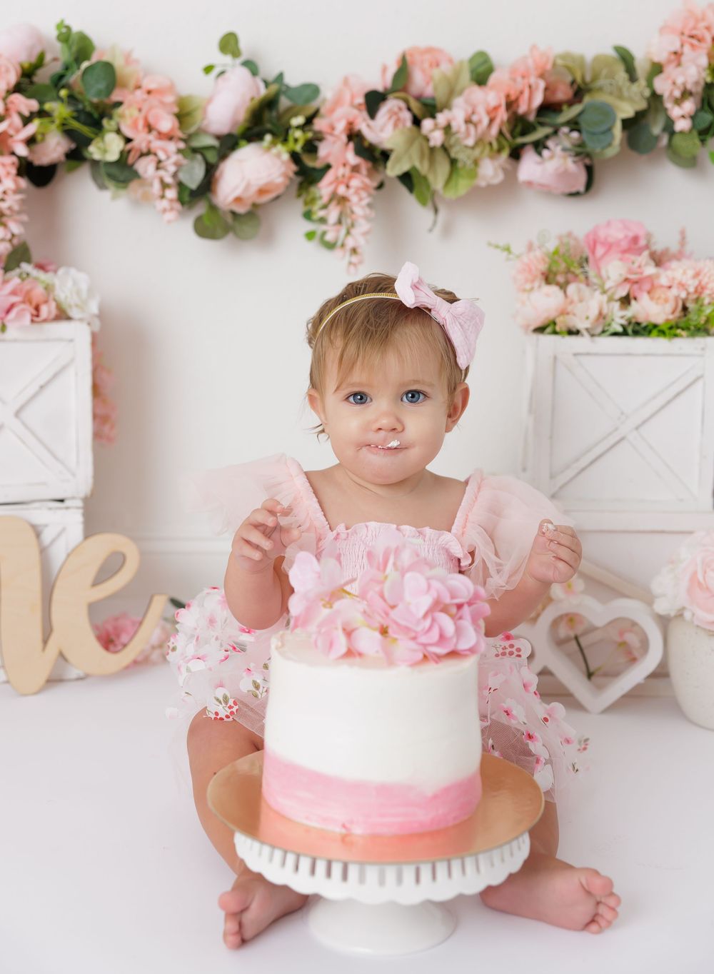 Baby girl in pink tutu smiles at first birthday cake smash with floral backdrop and pink roses.