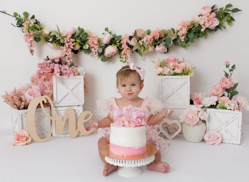 Baby girl in pink romper smashes first birthday cake surrounded by blush floral arrangements and decorative white boxes.