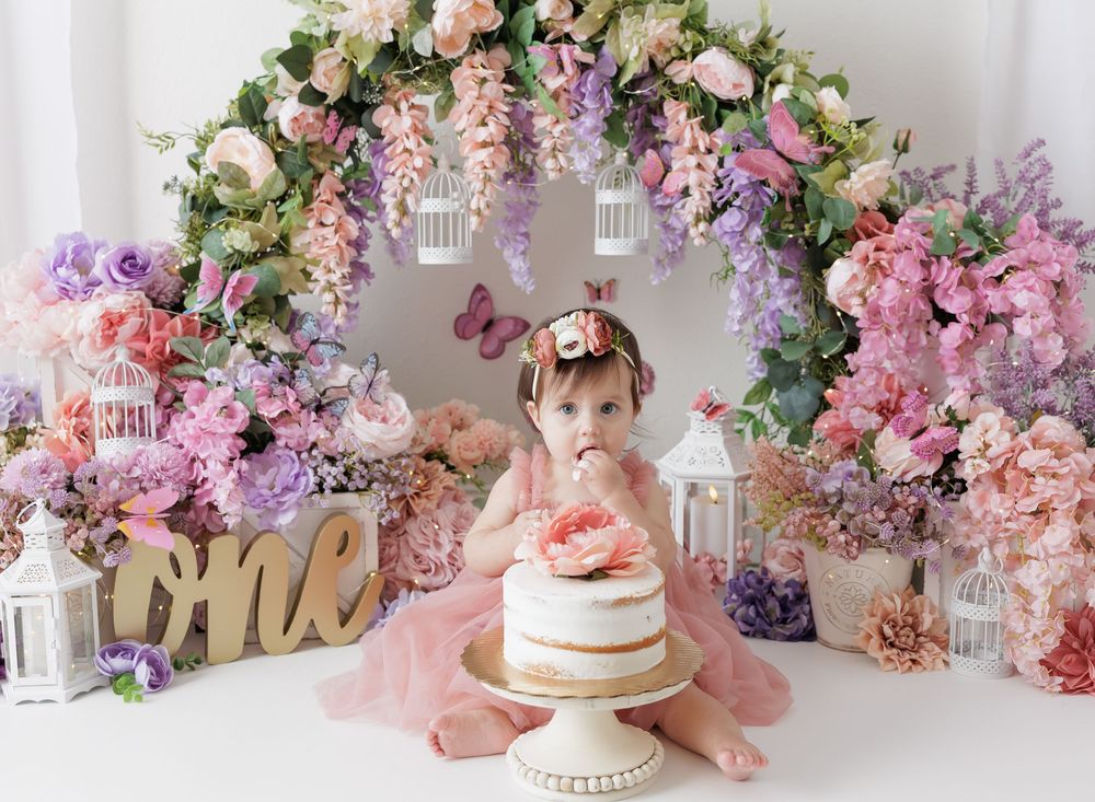 Baby girl in pink dress smashes first birthday cake surrounded by floral arch with birdcages and pastel flowers.