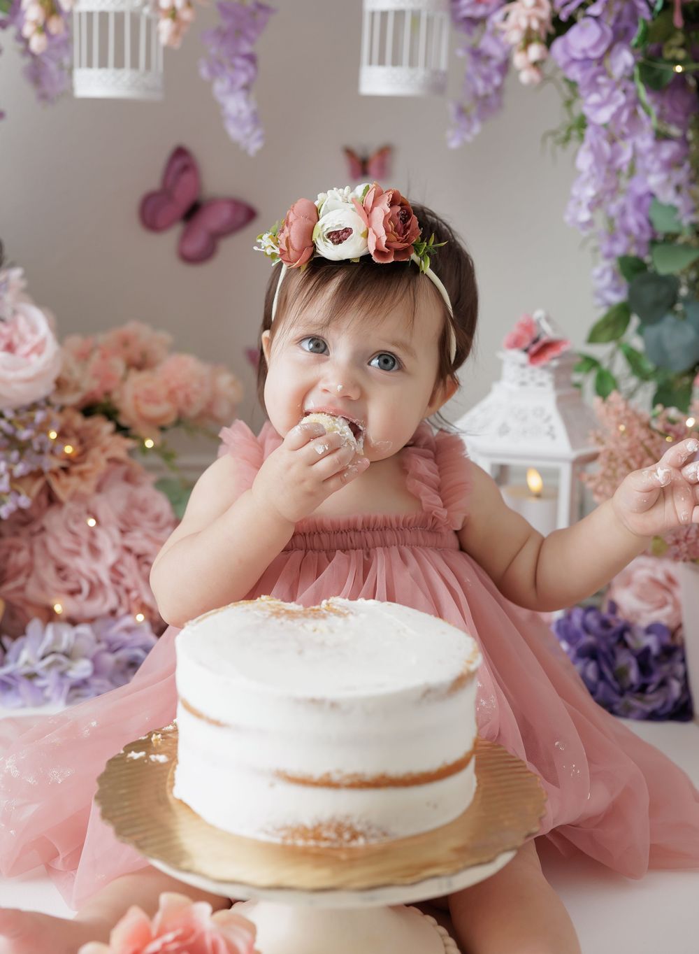 Baby girl in pink dress and floral crown smashes first birthday cake surrounded by pastel flowers.