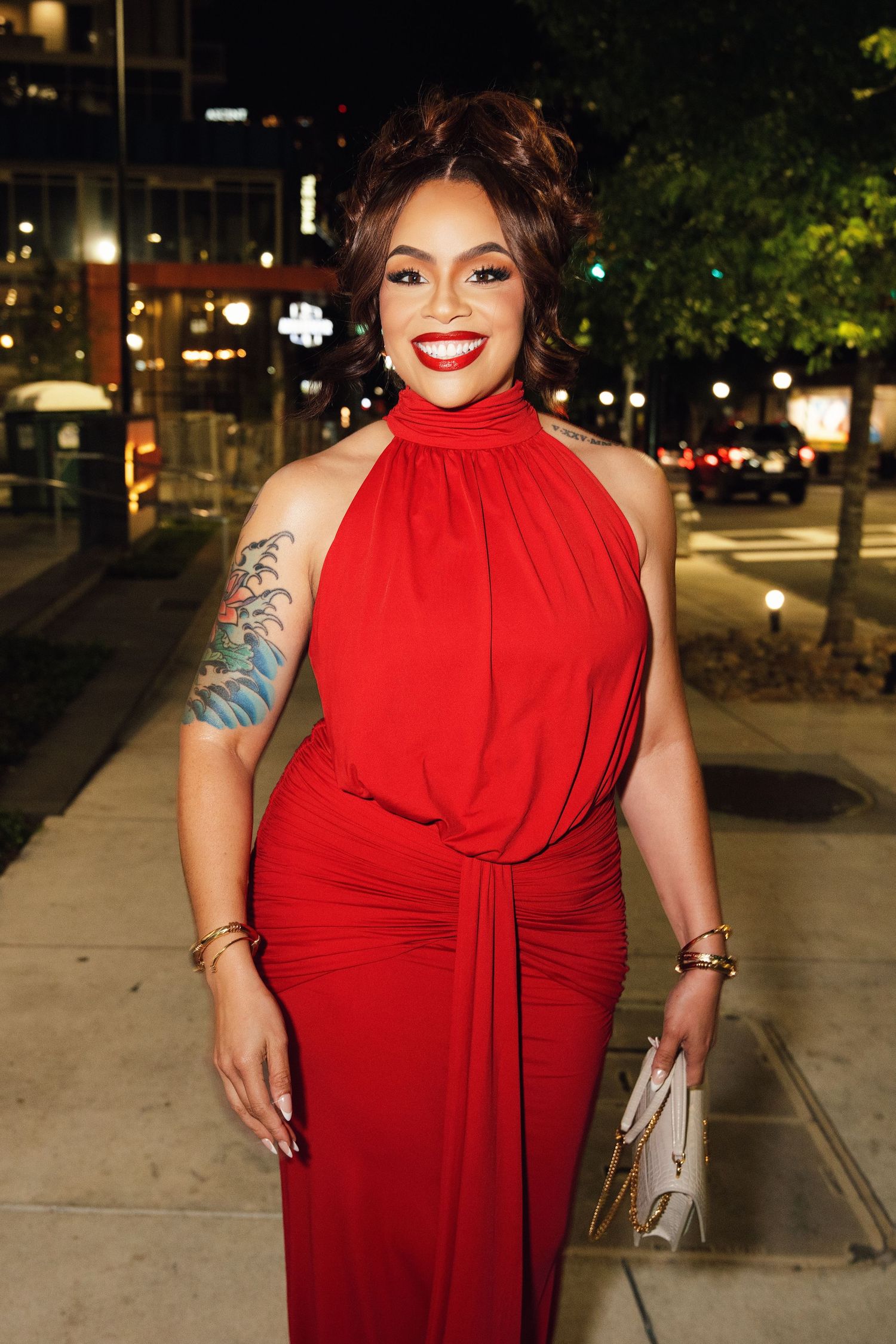 Woman in black sequin one-shoulder gown poses outdoors at night with golden bokeh lights in background.