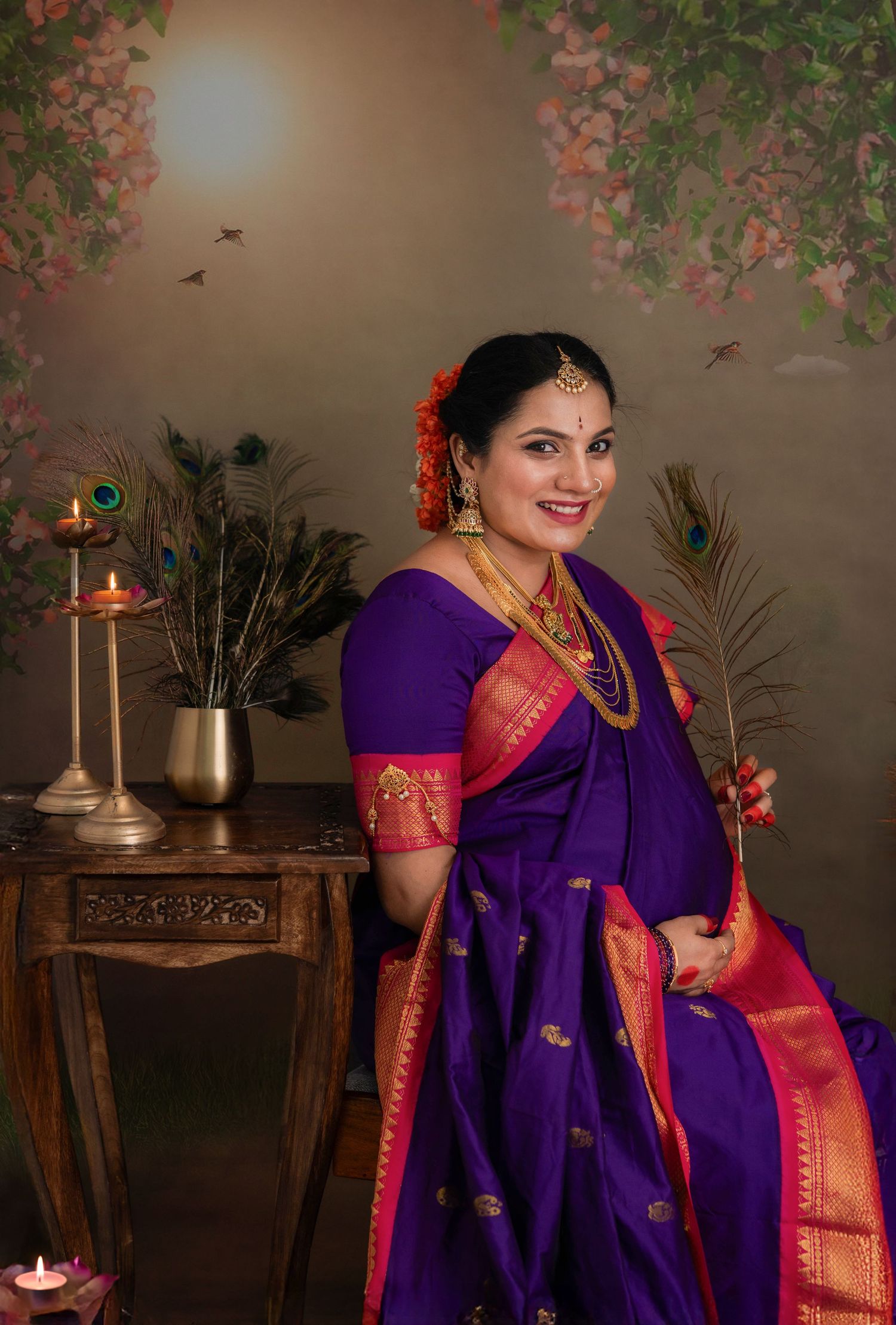 Expecting mother in a purple and pink silk saree holding a peacock feather, seated beside a wooden console with candles and floral decor