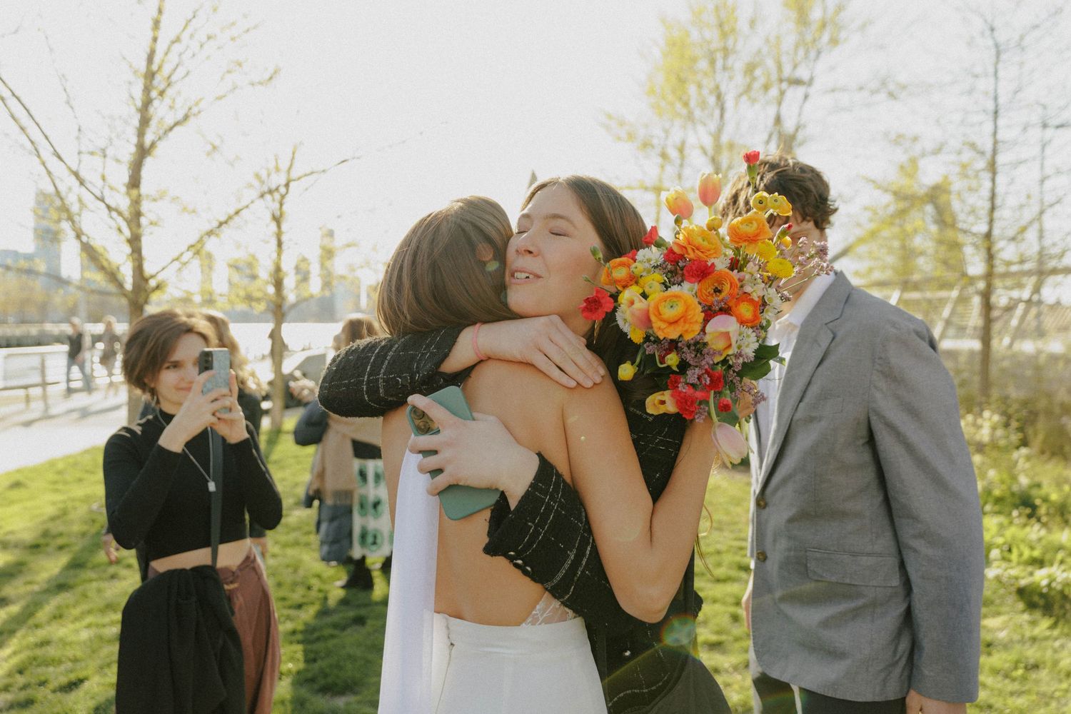 Bride in white dress hugs guest while holding colorful bouquet at outdoor wedding ceremony.