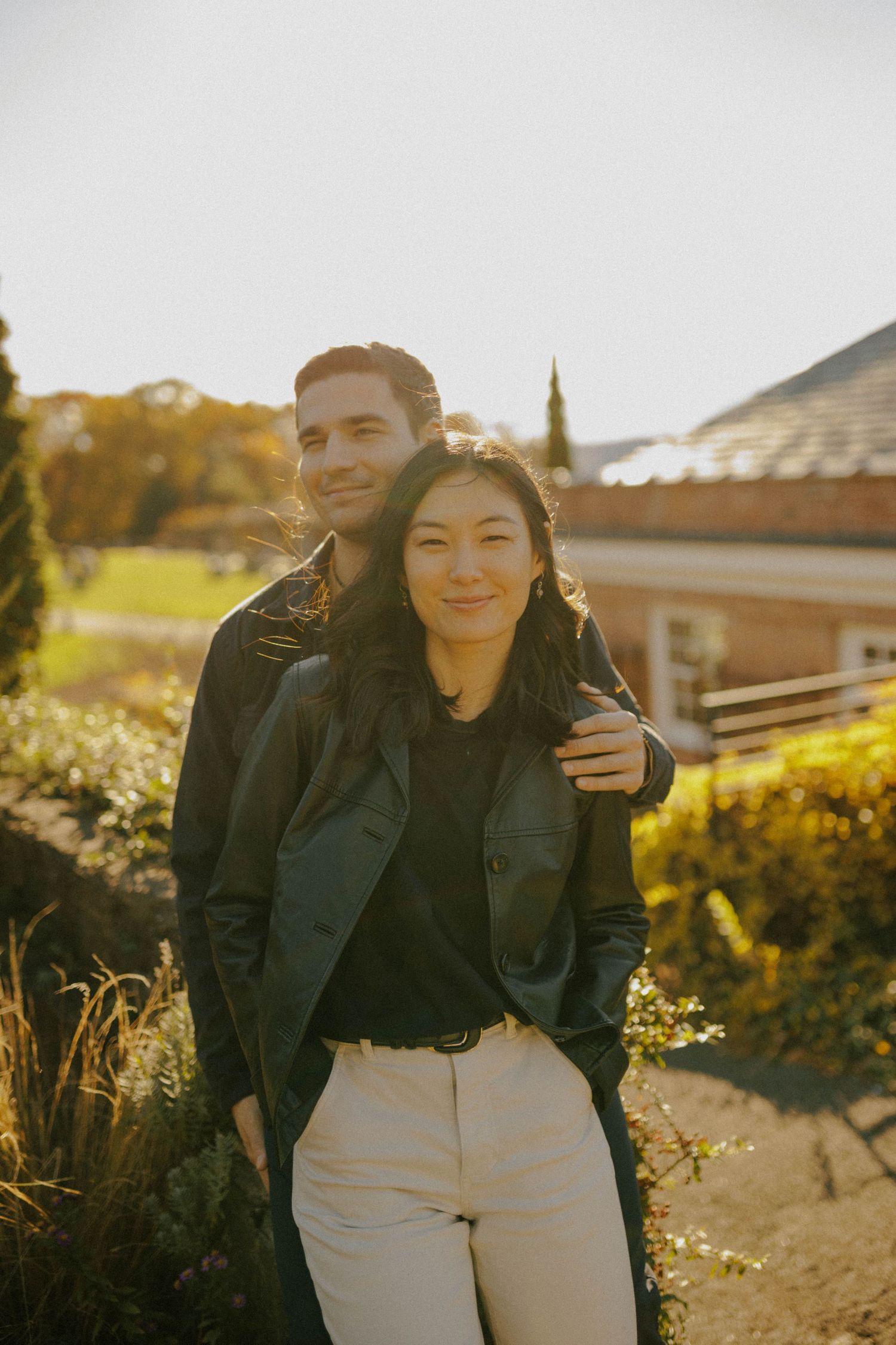 A couple poses outdoors in golden hour light, man in black jacket embracing woman in cream pants near a rustic building.