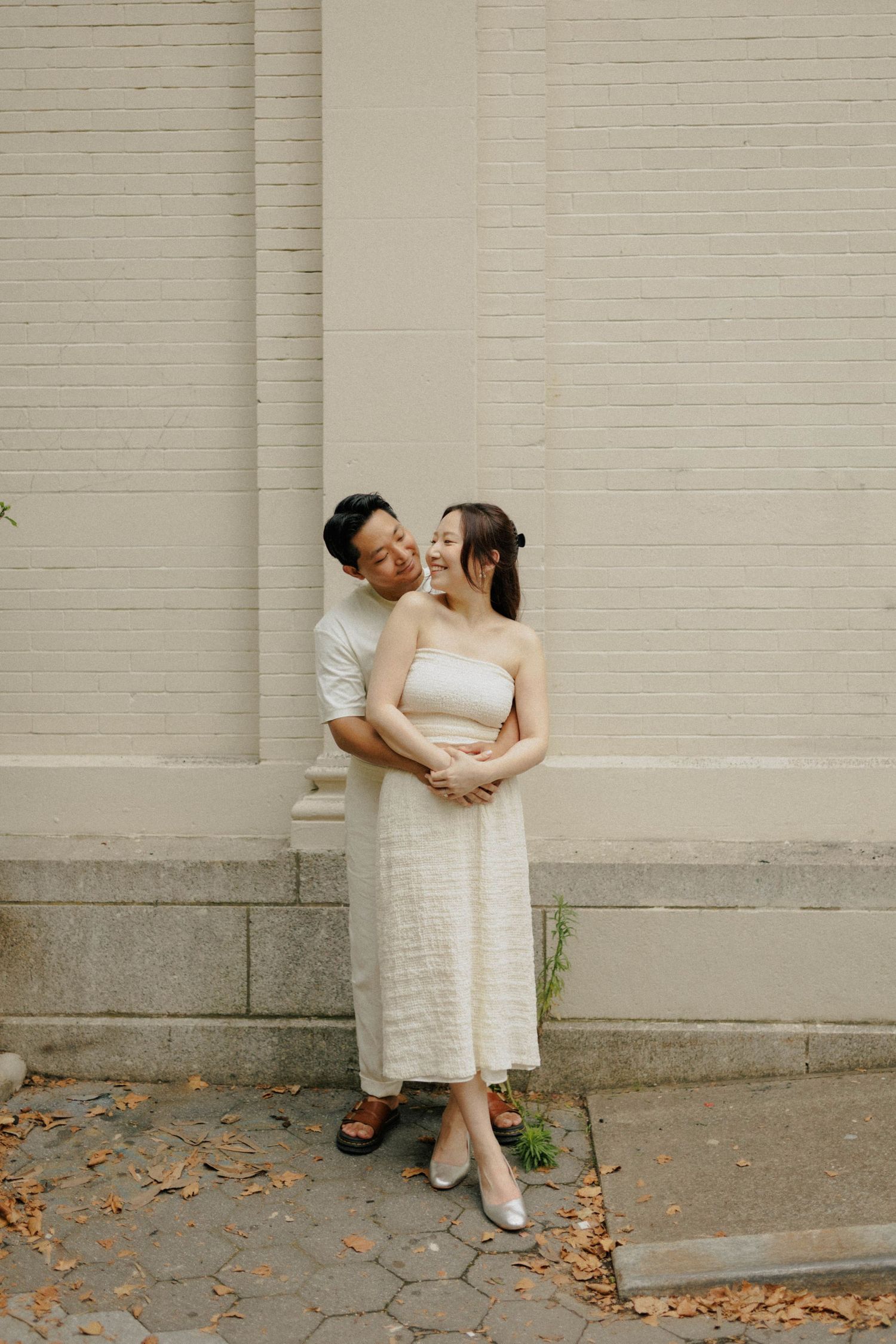 Couple embracing and smiling against a cream brick wall, wearing neutral tones, standing on a cobblestone path.
