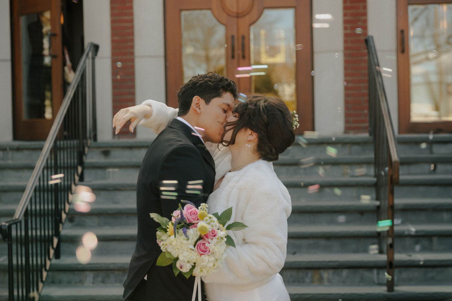Newlywed couple sharing a kiss on church steps, bride holding pink bouquet in white fur coat, groom in black suit.