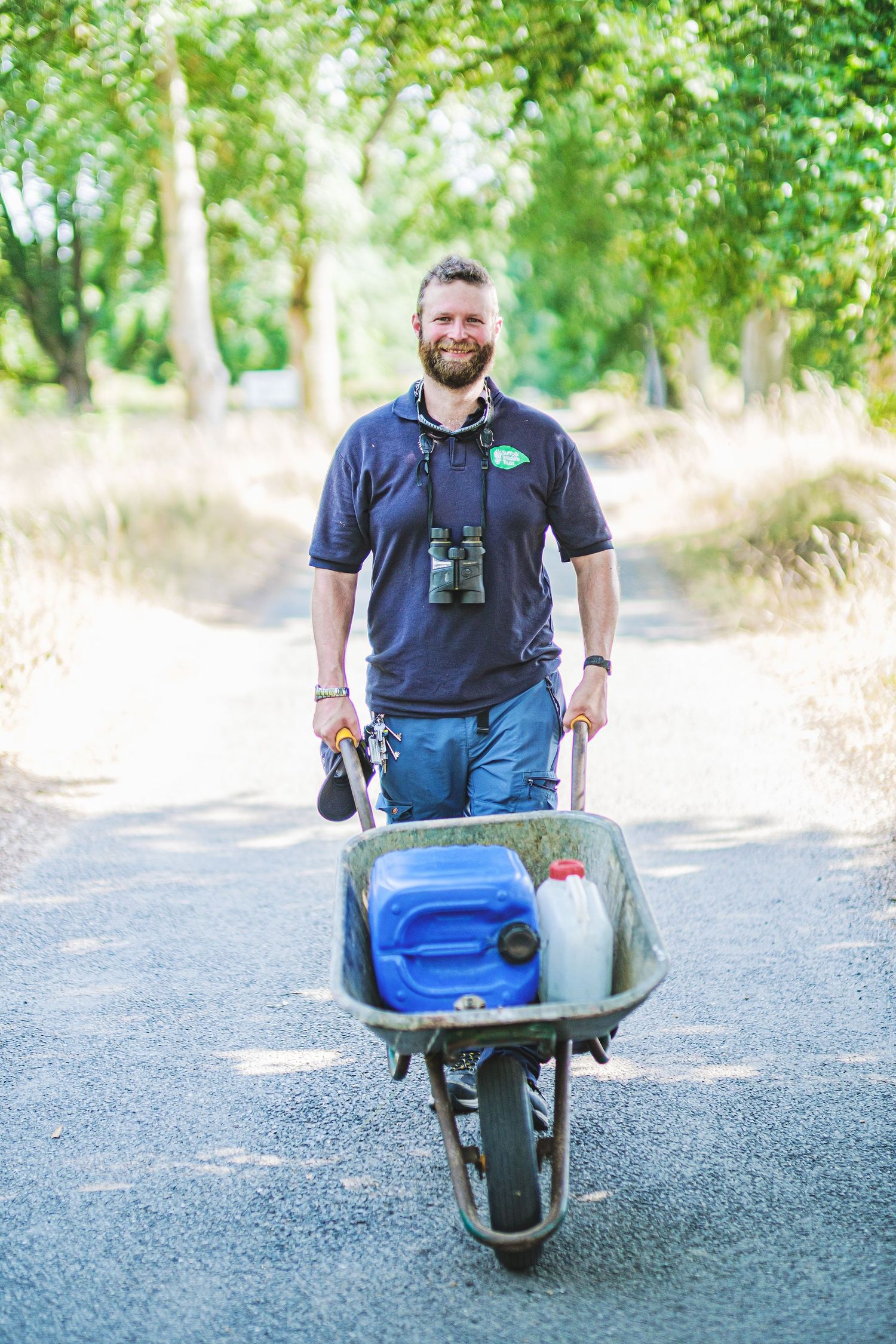 Suffolk Wildlife Trust: Smiling bearded man pushing a wheelbarrow with blue & white containers on a sunny, tree-lined path. By John Ferguson