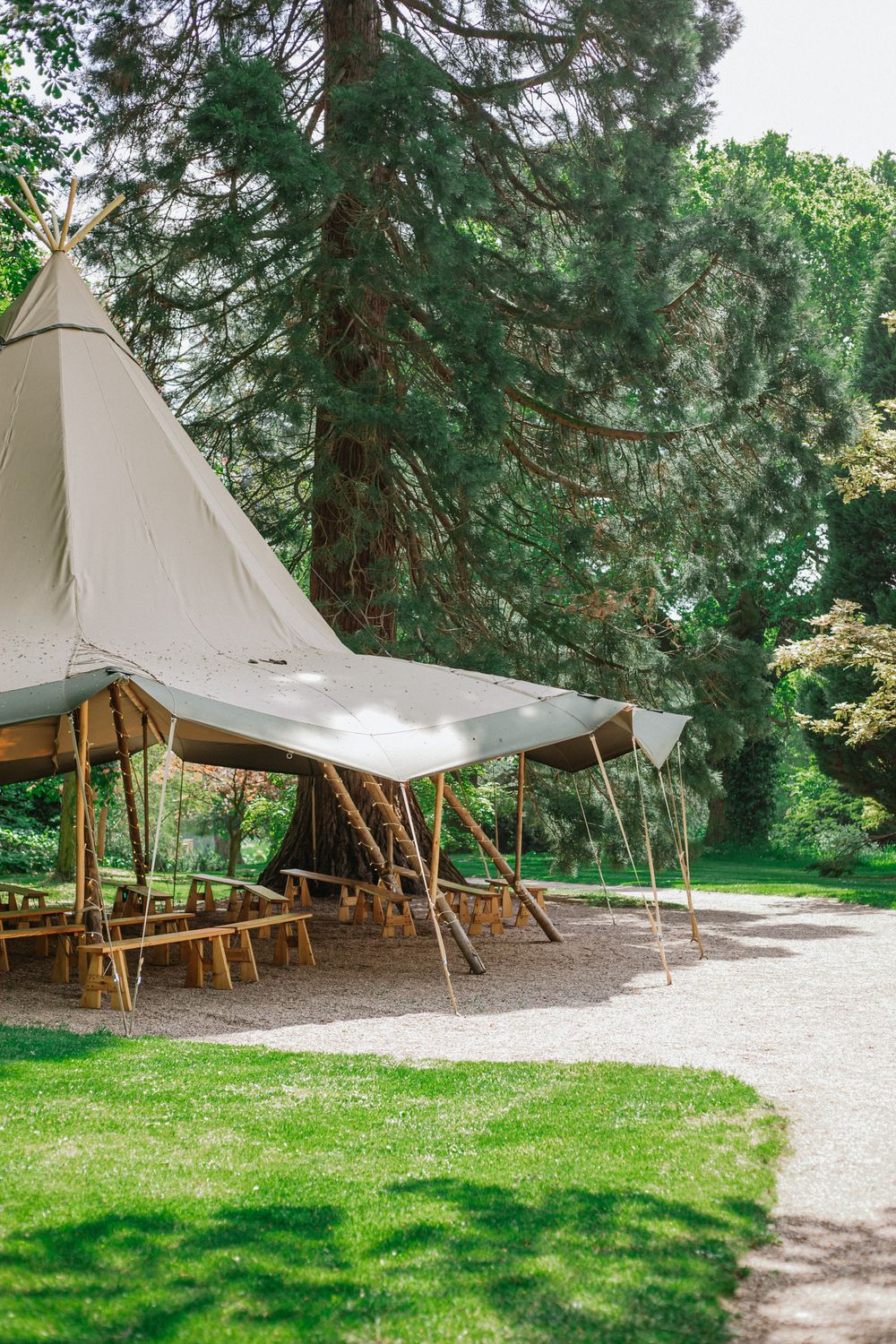 White canvas whatton tipi tent with wooden poles and picnic benches set up outdoors in a lush green garden setting.