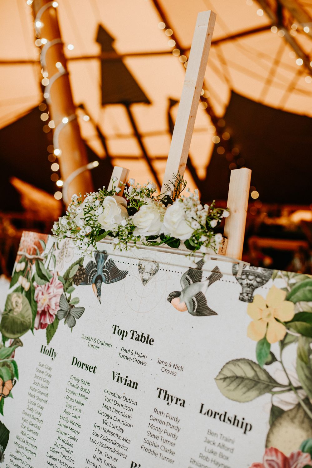 Floral wedding seating chart with white roses displayed inside a warmly lit whatton tipi venue with fairy lights.