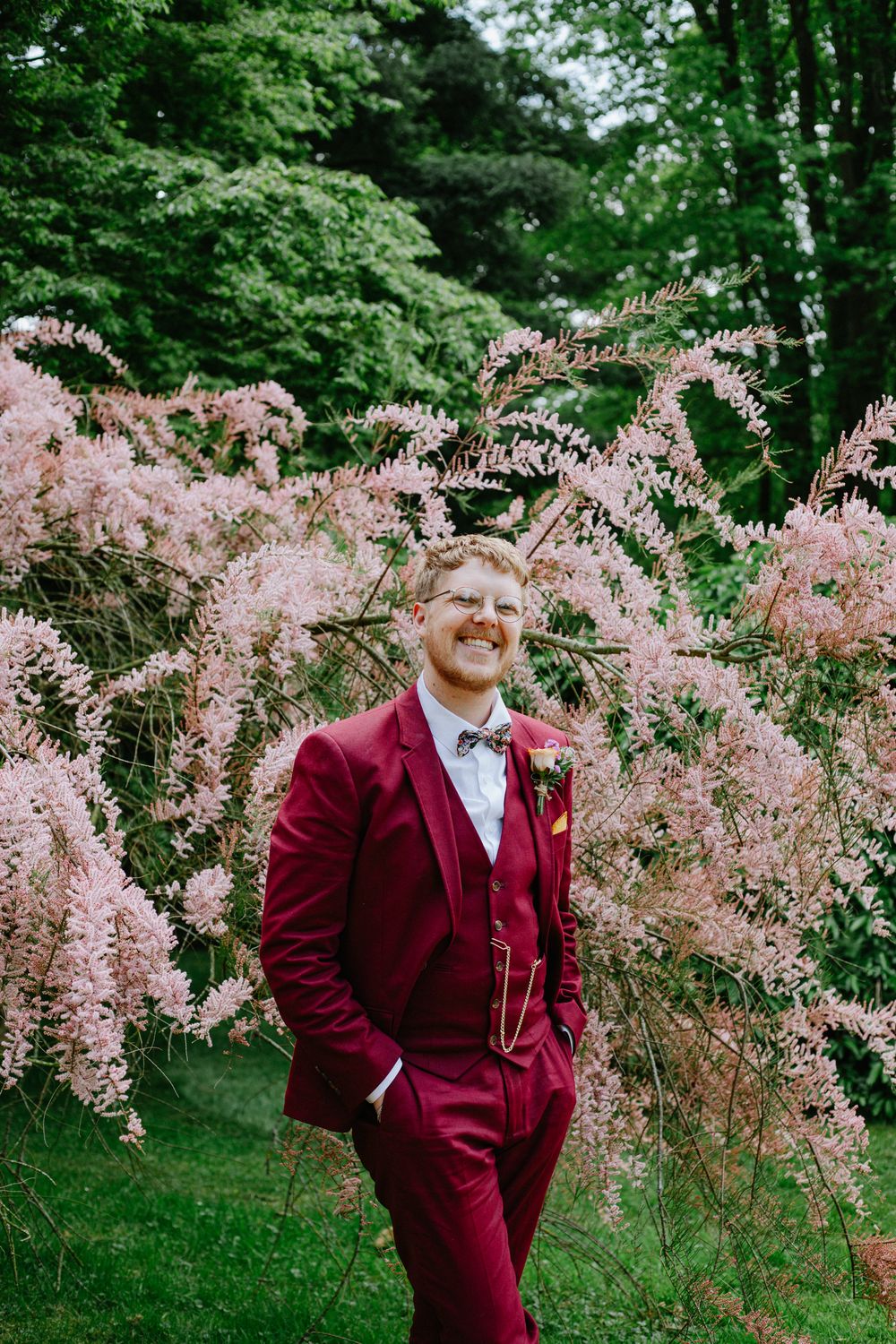 Smiling groom in burgundy suit and bow tie standing before pink flowering trees at Whatton House Tipi wedding venue.