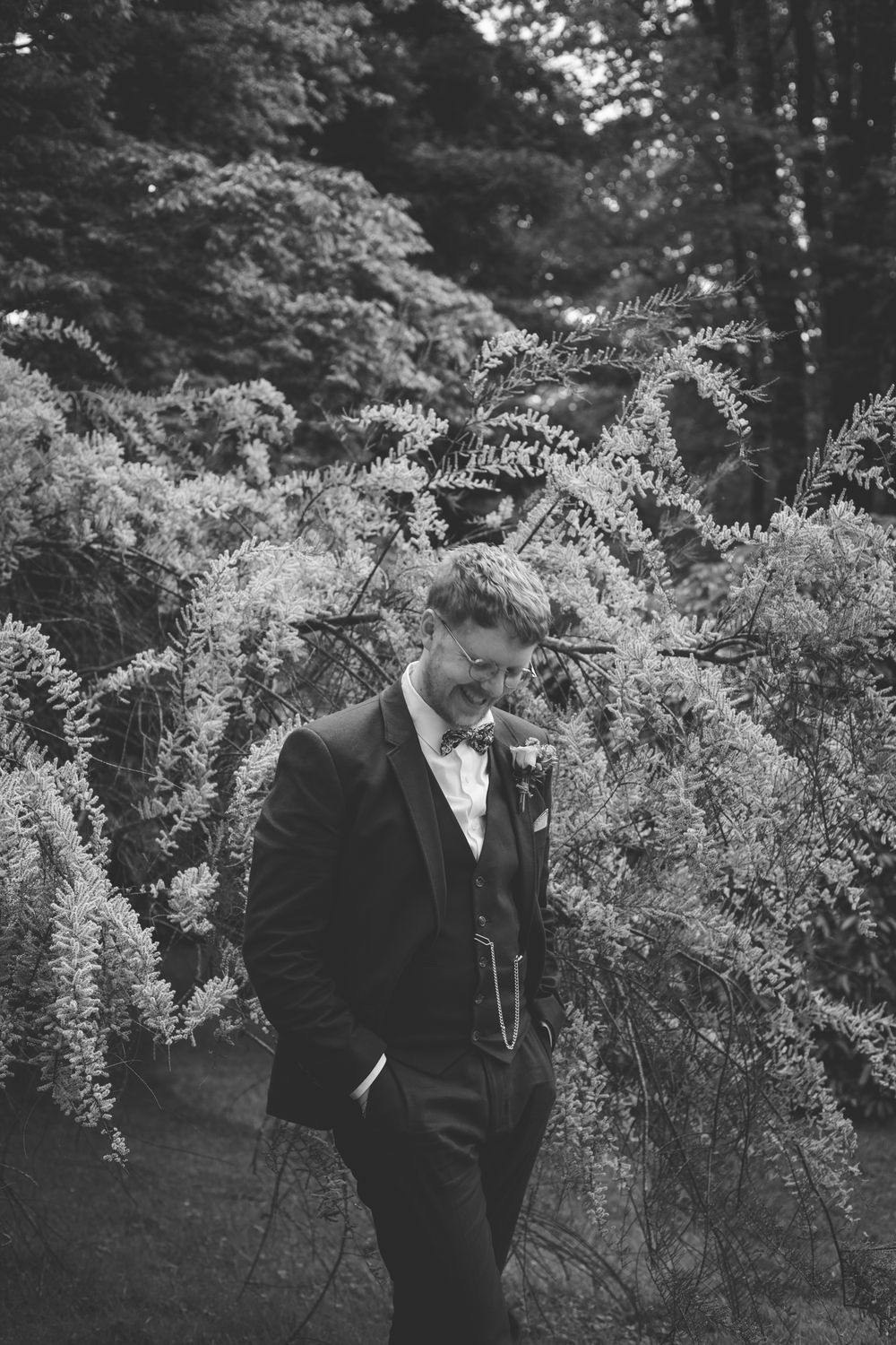 Groom in black suit smiling downward surrounded by lush trees in a black and white wedding photo.