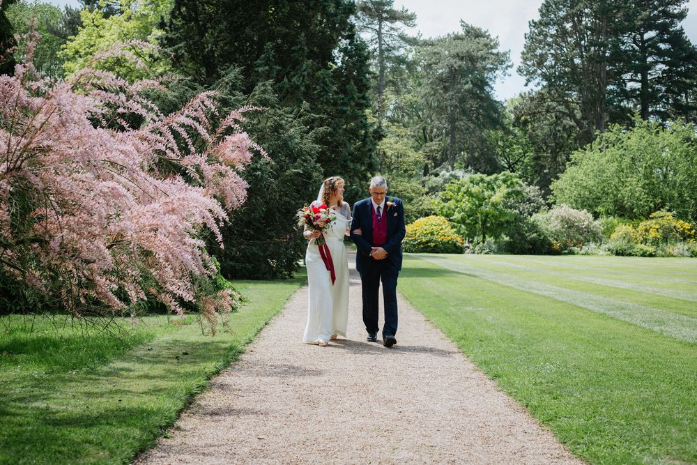 Bride and her dad walking hand in hand along a garden path surrounded by lush greenery and pink flowering trees.