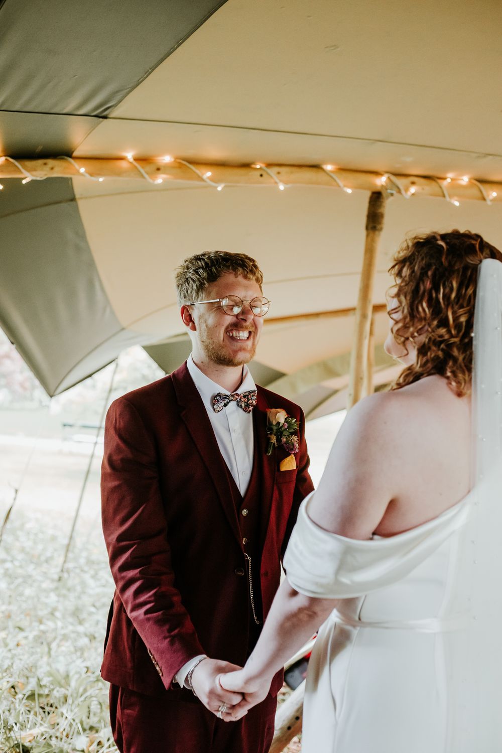 Groom in burgundy suit holds brides hands during wedding ceremony under a whatton tipi with fairy lights.