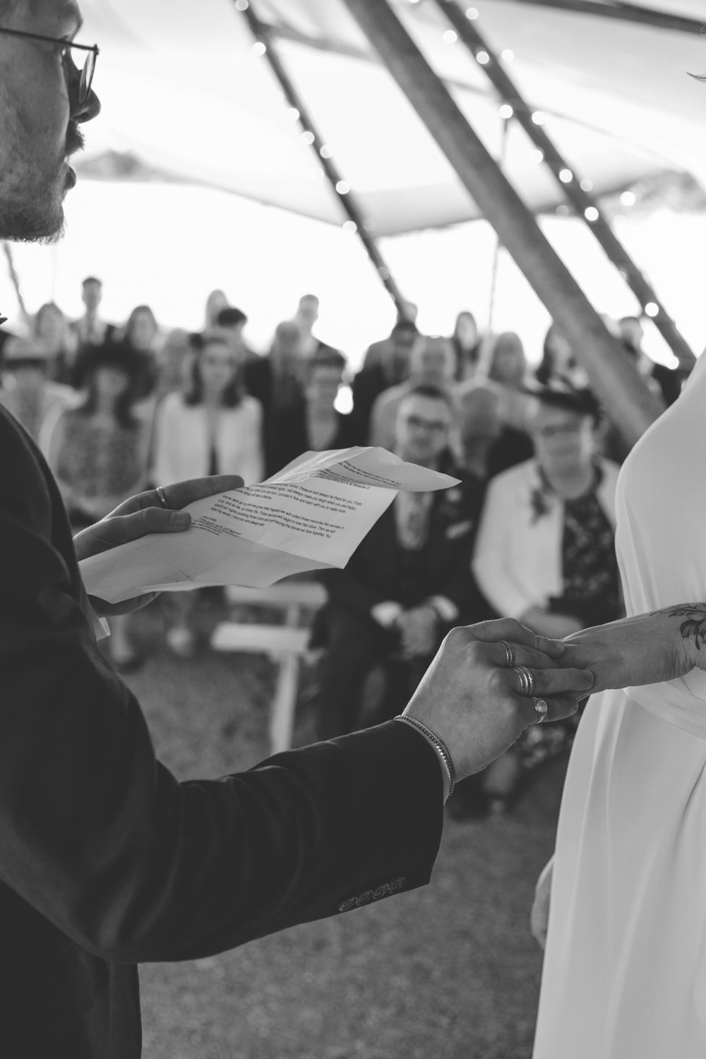 Groom reading wedding vows from paper while holding hands with bride during outdoor whatton tipi ceremony.