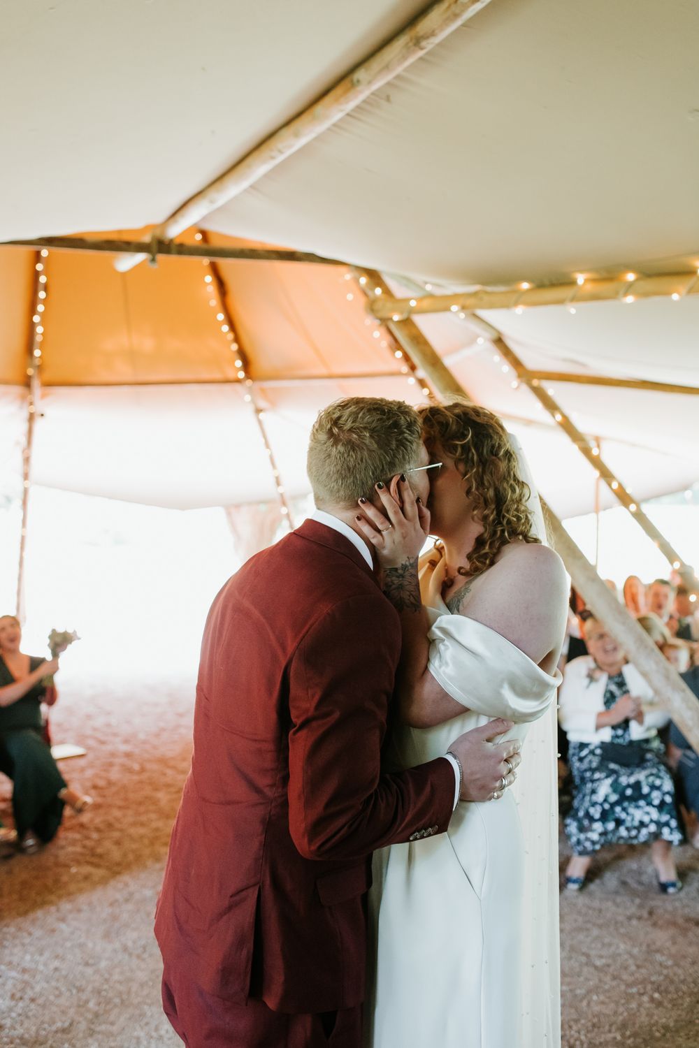 Bride and groom share first kiss under a rustic tipi tent decorated with fairy lights at their wedding ceremony.