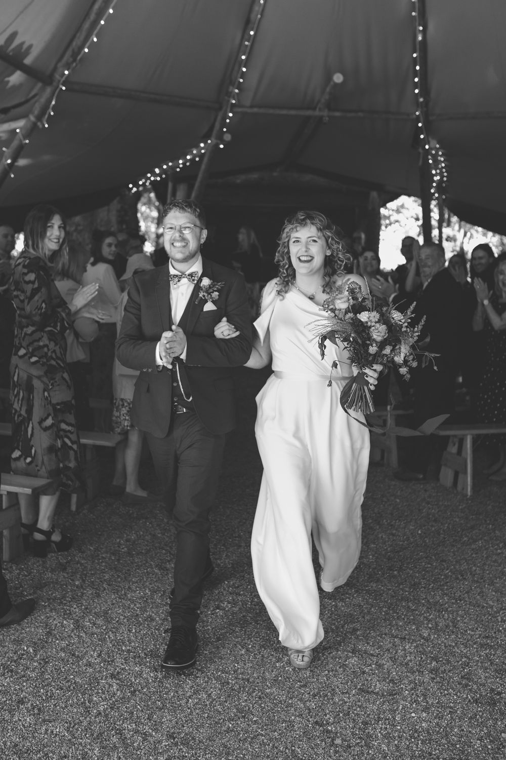 Black and white photo of a joyful bride and groom walking down the aisle under a tipi with string lights at their wedding.
