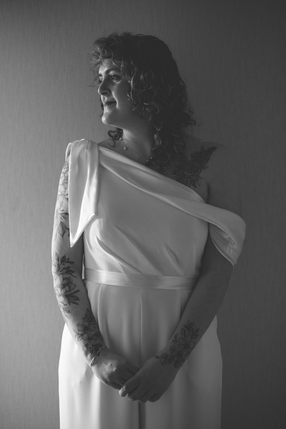Black and white portrait of a tattooed bride in an elegant one-shoulder white Davids Bridal Jumpsuit, gazing thoughtfully to the side.