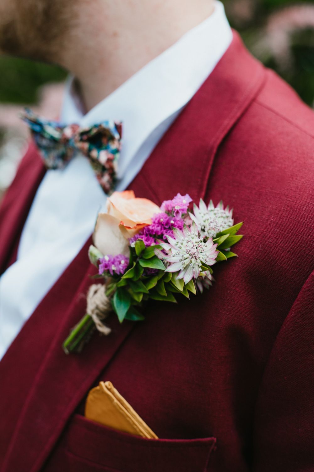 Groom in burgundy suit with floral boutonniere featuring peach rose and purple blooms, paired with a colorful bow tie.