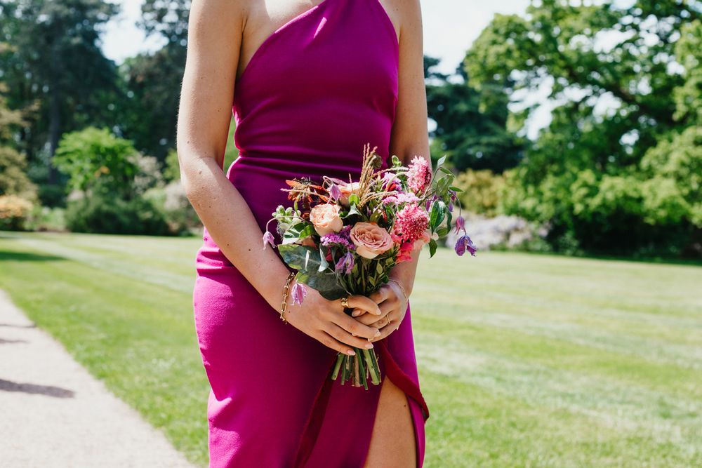 Bridesmaid in magenta one-shoulder gown holding a pink and purple floral bouquet outdoors.