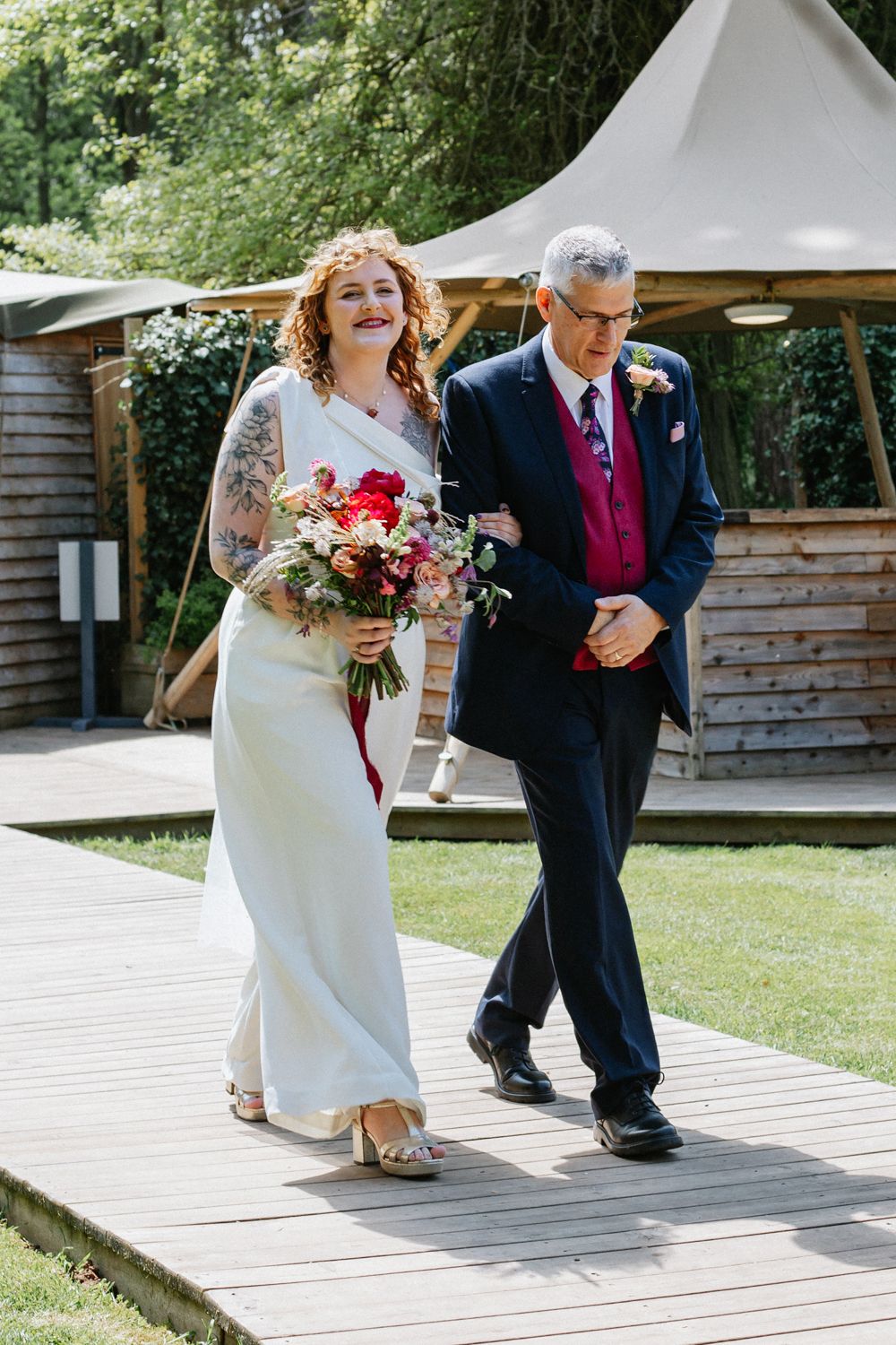 Bride with tattoos in white davids bridal jumpsuit holds red bouquet, escorted by her dad in navy suit on wooden path.