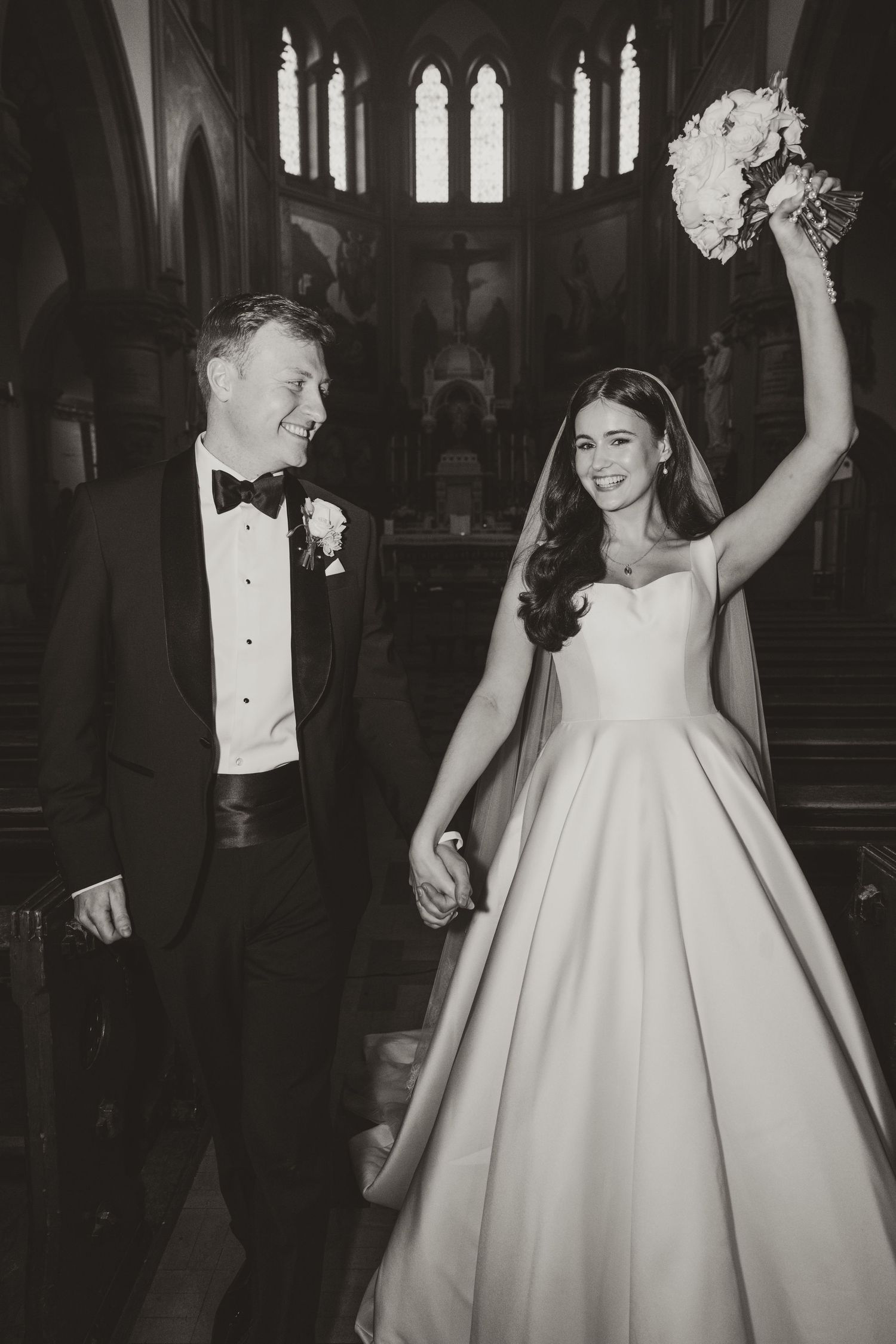 Black and white photo of a joyful bride and groom walking down church aisle, bride raising bouquet in celebration.