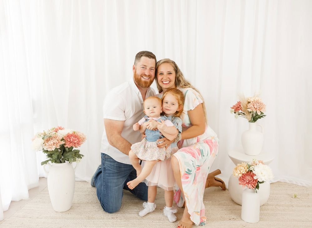 A smiling family of four poses together in a bright white studio with pink floral arrangements.