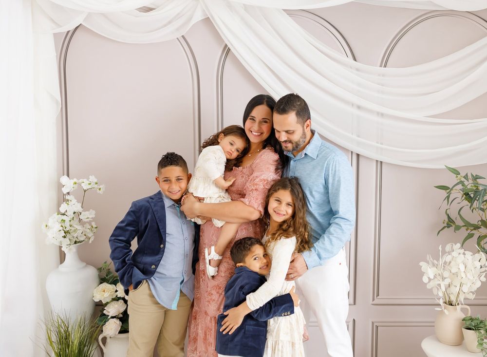 Family of six smiling together in an elegant white studio setting with floral decorations and draped fabric.