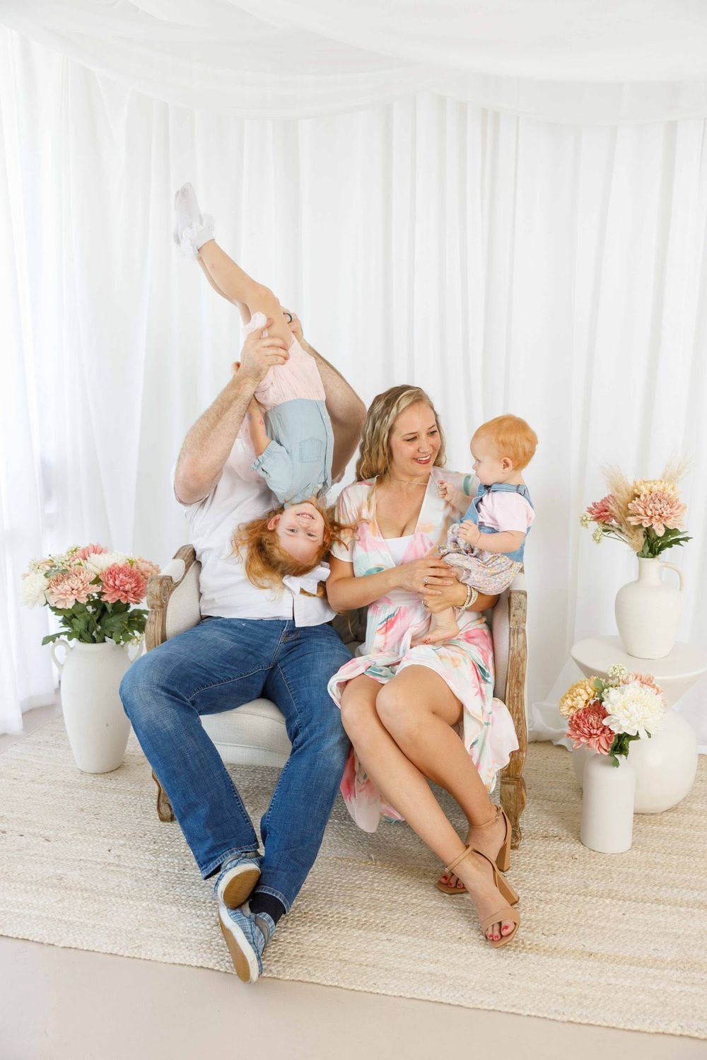 Happy family with two young children playing and laughing together in a bright white room with floral arrangements.