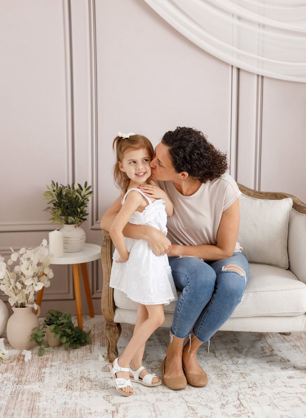 A mother kisses her young daughter on the cheek while sitting on a couch in an elegant, softly lit room.
