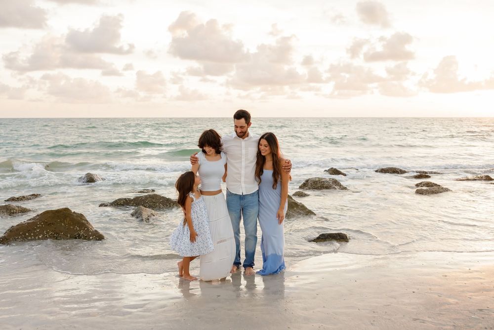 Family of four posing together on a sandy beach at sunset with ocean waves and rocks in the background.