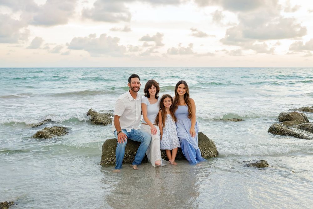 Family of four sitting on ocean rocks at sunset, wearing coordinated blue and white outfits at the beach.