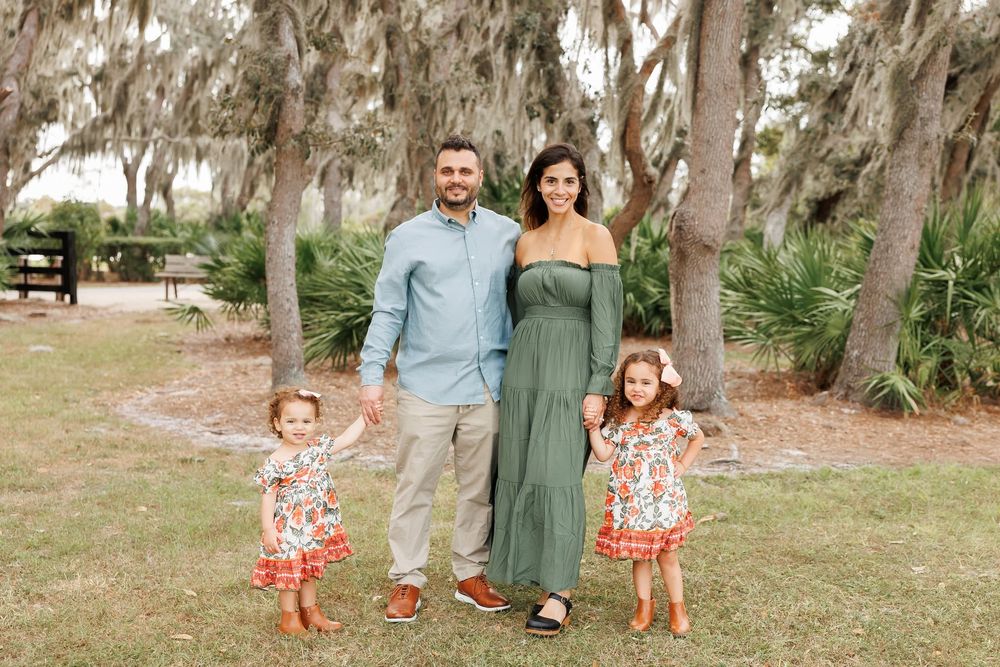 Young couple with two toddler girls holding hands outdoors under Spanish moss trees.