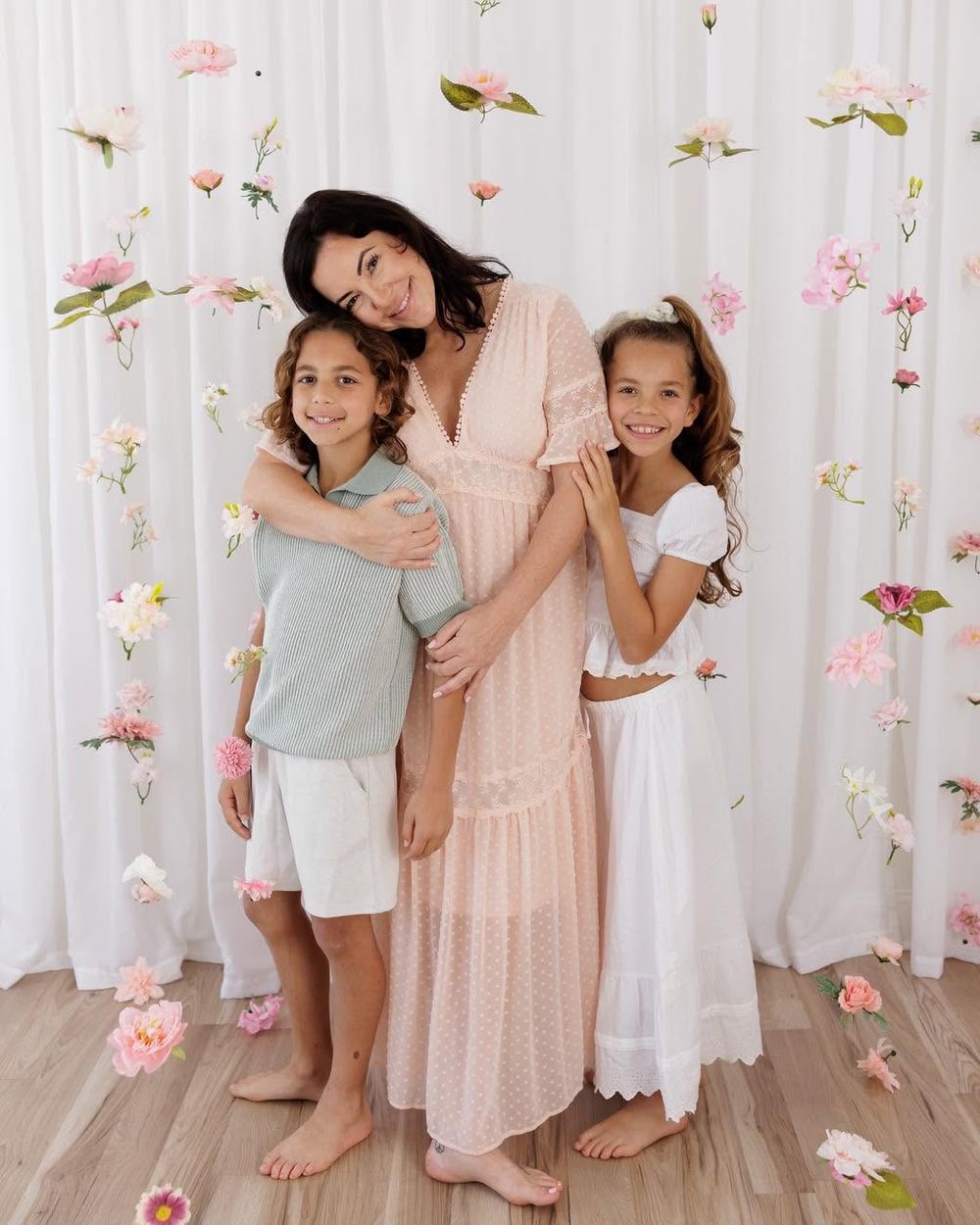 A woman in a pink maxi dress poses with two children against a floral backdrop with hanging flowers.